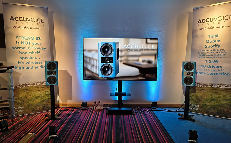A pair of AccuVoice Stream S3 in a demonstration room at a HiFi Show.  There's a screen with a picture of the speaker on it.  The speakers are a blue colour.