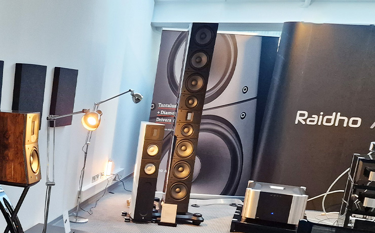 the Raidho TD6 floorstanding loudspeaker beside a white Raidho X2.6 loudspeaker with a Moon  power amp on a low stand the other side.  there are lamps and a Raidho backdrop in the photo too along with a bookshelf speaker on a speaker stand.  The photo was taken at the Munich 2024 HiFi Show