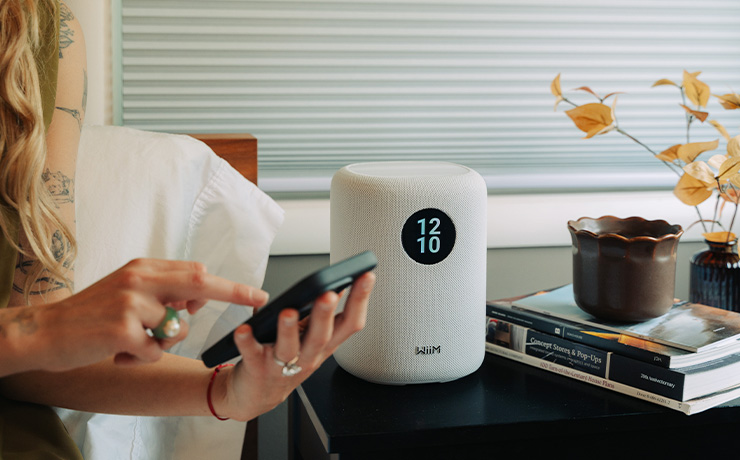 A white WiiM Sound speaker on a black table beside a stack of books.  A woman is sitting beside it holding a smartphone