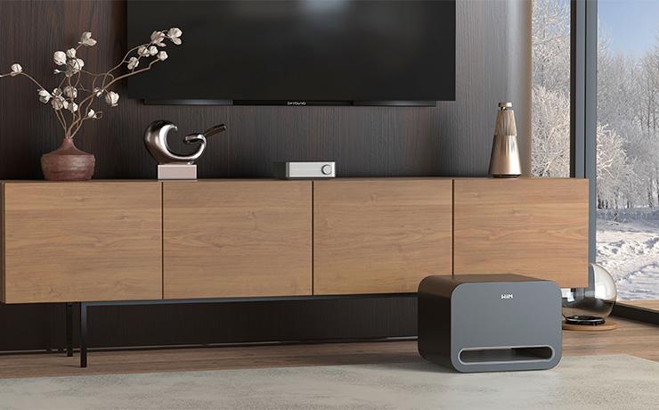 The WiiM Sub Pro in the black finish on a floor in front of a long wooden sideboard. There's a large window to the right with snowy woods visible