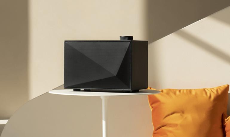 An Astell & Kern Speaker on a table with a partial view of an orange cushion behind.