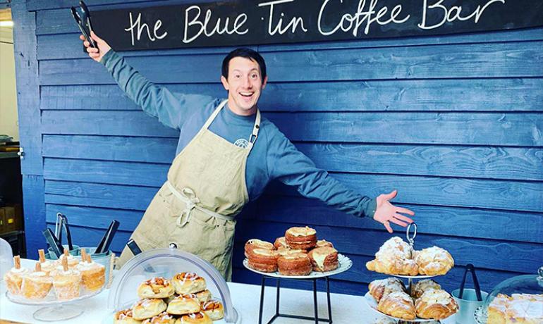 Image shows a very happy man with his arms outstretched standing in front of a blue wooden shed.  In front of him is a table laden with cakes.