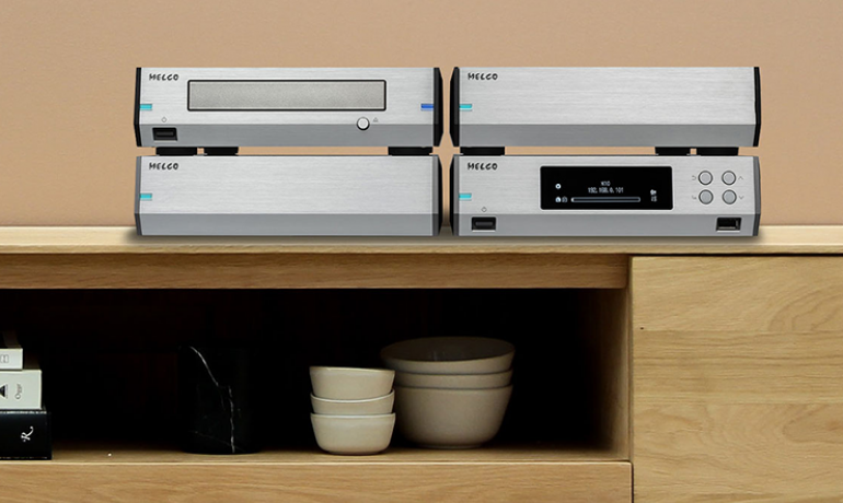 Four melco products on top of a wooden sideboard with bowls on a shelf underneath.  The products include the Melco D100 CD drive and the Melco N100 HiRes Music Library.  The wall behind is a light brown colour.