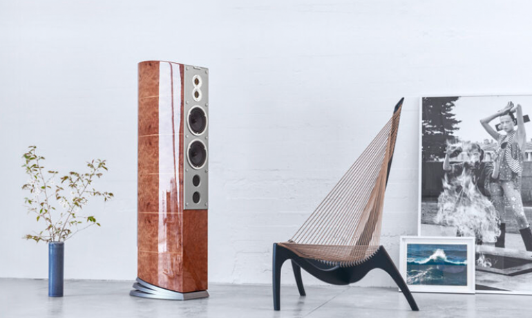 A tall Audiovector speaker angled to the right of the image.  The speaker is on the left of the image with a vase of twigs beside it on the left and, to the right, an artistic chair with pictures propped against the wall.