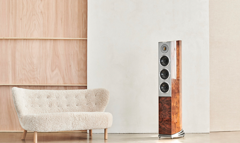 A tall Audiovector R8 speaker to the right of an image with a cream sofa on the left.  There's a natural wood coloured wall behind to the left and a painted wall behind to the right.  The painted wall is cream.  The floor is shiny and a beige colour.