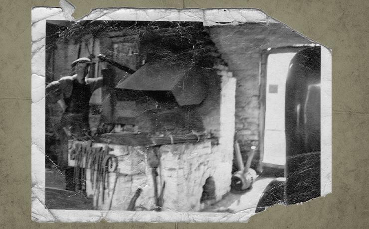 An old, creased black and white photograph of a blacksmith inside a forge leaning against the equipment