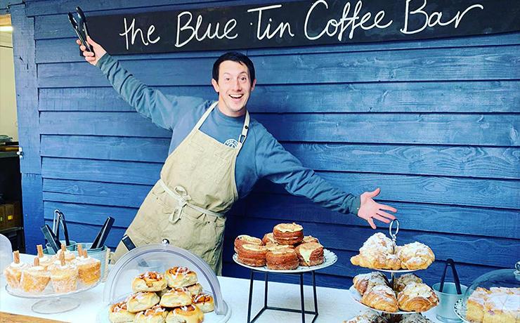 A very happy man in front of a blue wooden structure.  In front of him is a table with some lovely looking cakes and pastries.