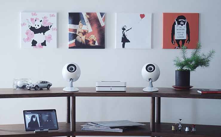 A pair of Eclipse TD307 speakers in white on a shelving unit with an iPad on a stand, a plant and some other objects.  there are four canvas pictures on the wall behind the shelves.
