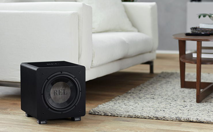 REL HT/1003 Subwoofer on a wooden floor to the side of a white sofa.  There's a grey rug on the floor and a brown wooden coffee table on the rug.  The walls are grey.