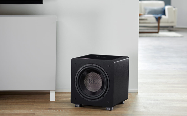 REL HT/1205 Subwoofer in black on a laminate floor in front of a slim cabinet.  In the background is a sofa.
