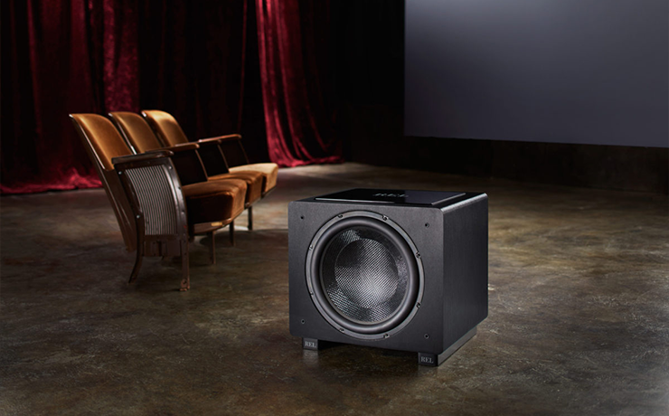 REL HT/1508 Predator Subwoofer on a brown floor with red velvet curtains to the left and one row of old theatre seats.  There is what looks like a cinema screen in the background.