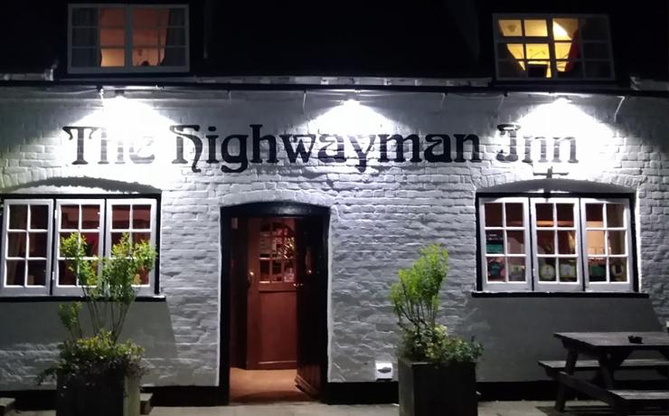 The front of the Highwayman Inn which is white painted brick.  The photo was taken at night and there are lights shining on the name.  There's a door in the centre with a window either side.