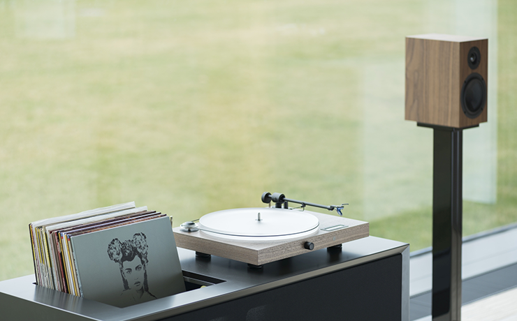 Project Juke Box S2 turntable on a wooden unit containing vinyl in front of a window with the Project loudspeaker beside it.