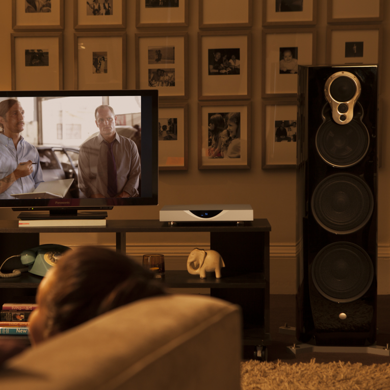 A Linn Klimax 350 loudspeaker beside a television stand with a tv on and a Linn Klimax DSM.  There's a wall of black and white photographs behind and a person sitting on the sofa in the foreground.