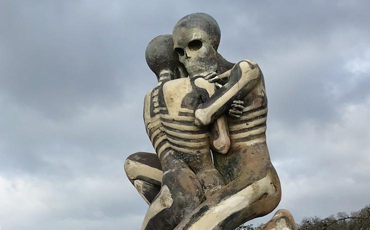 A large sculpture of two people embracing against the sky.  They are grey with white painting showing the bones.
