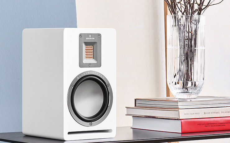 Audiovector QR1 Loudspeaker in white on top of a table.  Beside the speaker is a stack of three books on top of which is a vase of twigs.