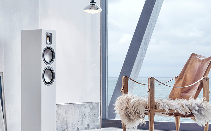 An Audiovector QR3 Loudspeaker in white in a room with a sea view.  In front of the speaker is a canvas chair with a shaggy rug on it.