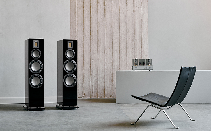 A pair of Audiovector QR 7 SE speakers in a minimalist space with a single chair on the right of the image