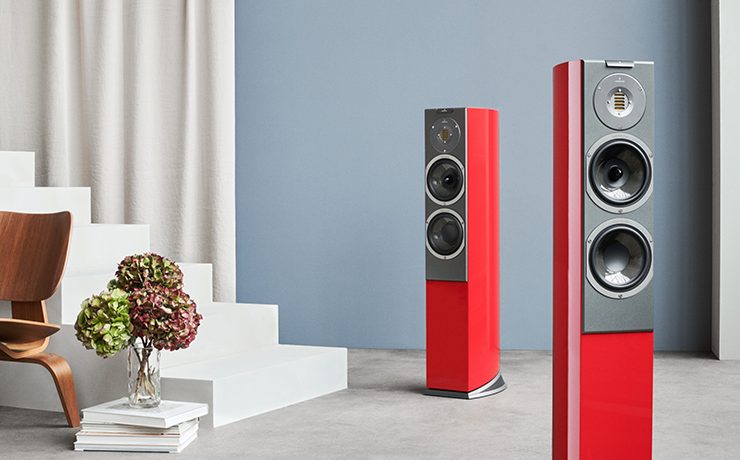 A pair of red Audiovector R3 Avantgarde Loudspeakers.  One in the foreground.  White steps on the left of the image, a wooden chair in front of the steps on the left and only partially visible.  The room has a grey/blue wall with a cream curtain on the left.  There's a vase on top of a stack of books with three dried hydrangea blooms.