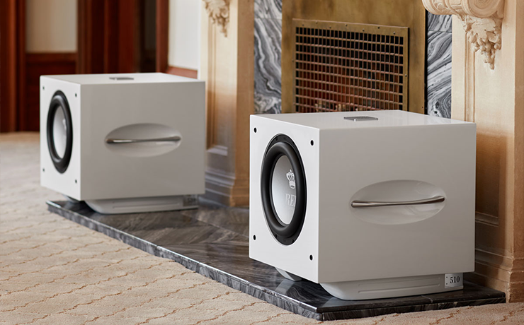 A pair of REL S/510 Subwoofers in white.  They are either side of a fireplace that has a metal grille.  The fireplace is large.  The woofers are standing on a marble style tiled area.  The carpet is beige.