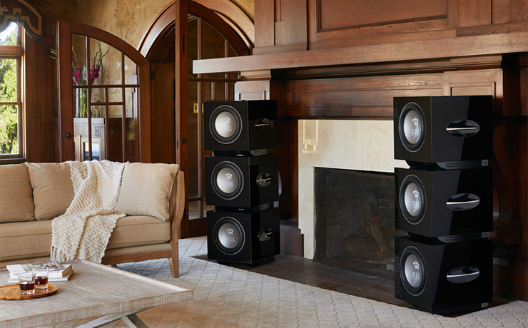 A stack of three REL S812 Subwoofers on either side of a fireplace in a large, high ceiling living room.  There are large wooden arch shaped double doors on the left of the fireplace.  There's a cream sofa with a wooden coffee table in front of it.  The floor is a beige carpet.