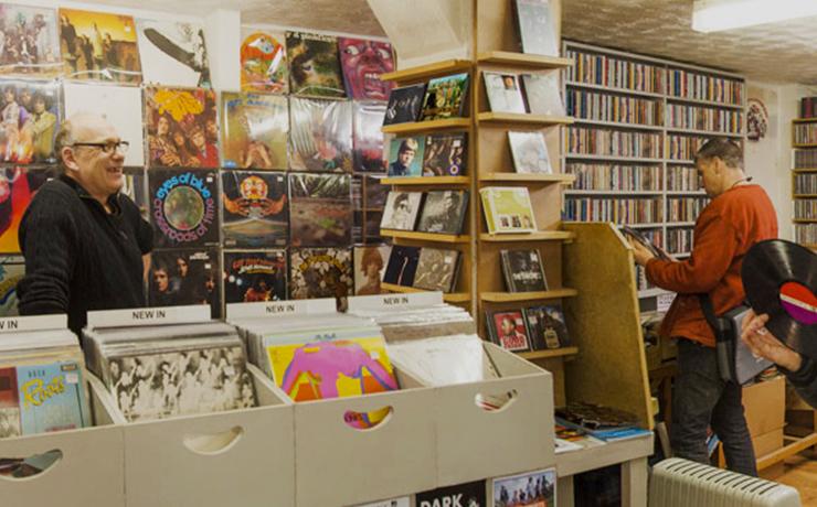 A picture of the inside of a record shop.  There are bins full of vinyl albums in the foreground with records lining the wall behind.  Between the wall and the bins, there's a middle aged man smiling.  he has glasses.  On the right, there are shelves of CDs on the wall with a man in a rust coloured jumper looking at what's there.  On the right of the image is a person's hand holding a vinyl record.