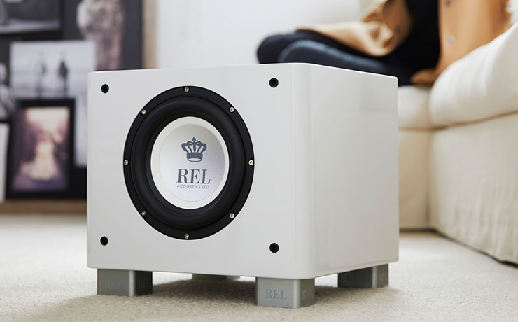 REL T/7x Sub-woofer in white on a cream carpet.  There's a cream sofa on the right with a lady seated on it.  In the background there's a selection of stacked images out of focus.
