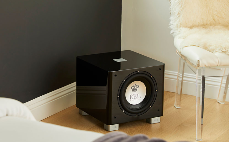 REL Acoustics T9x subwoofer.  The woofer is in the middle of the image.  To the right is a clear chair with a furry cover.  There's a bed partially visible in the foreground.  The floor is laminate.
