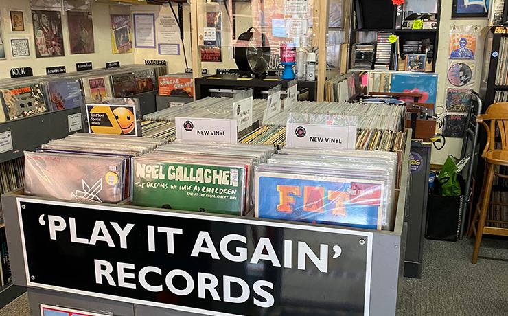 Image of the inside of the record store.  There are bins with vinyl in and a large sign saying "Play it Again Records".  there are posters on the sloped ceiling too.
