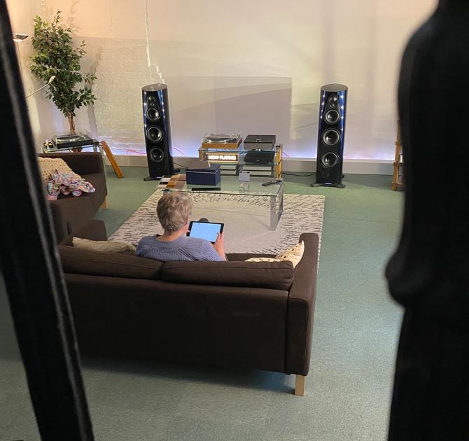 The ripcaster 360 speakers in Linn Heritage which is a dark blue colour.  There's a woman sitting on a brown sofa holding an iPad with her back to us.  Other HiFi equipment is between the two speakers.