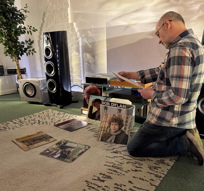 A man kneeling while reading the sleeve notes of an album.  there are Bob Dylan albums on the floor and stacked.  to the left of the image is the ripcaster 360 speaker in the Heritage colour.