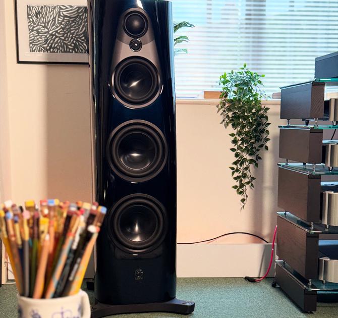 Linn 360 floor-standing speaker in Linn Heritage dark navy gloss finish, featuring multiple high-performance drivers for precise, dynamic sound. Shown in a modern listening room with a stack of hi-fi components, trailing green houseplant, and a ceramic mug filled with colourful pencils.