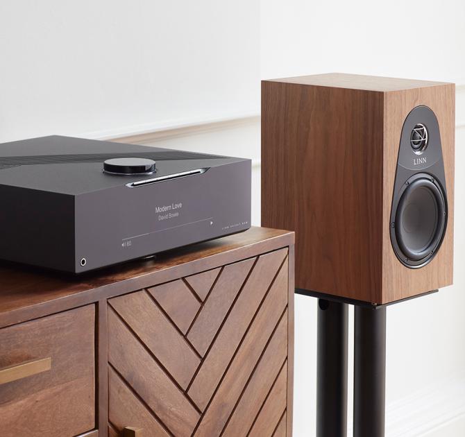 Linn 119 Loudspeaker in walnut on the black stand beside a wooden sideboard with a DSM on top of it.