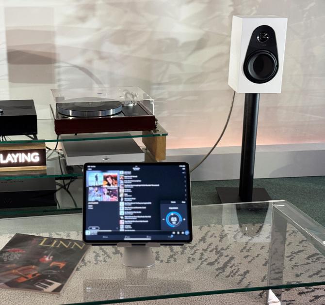 A white Linn 119 speaker at the ripcaster showroom.  It's on a stand beside an LP12 with an iPad in the foreground showing what's playing.