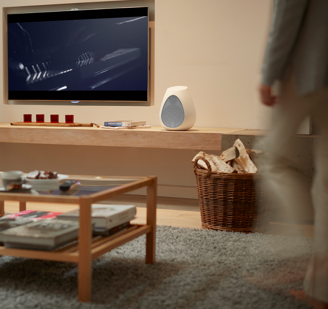 Linn Series 3 302 Loudspeaker on a side-board in a living room beside a television with a coffee table in the foreground and a blurred partial image of a person walking into the room.