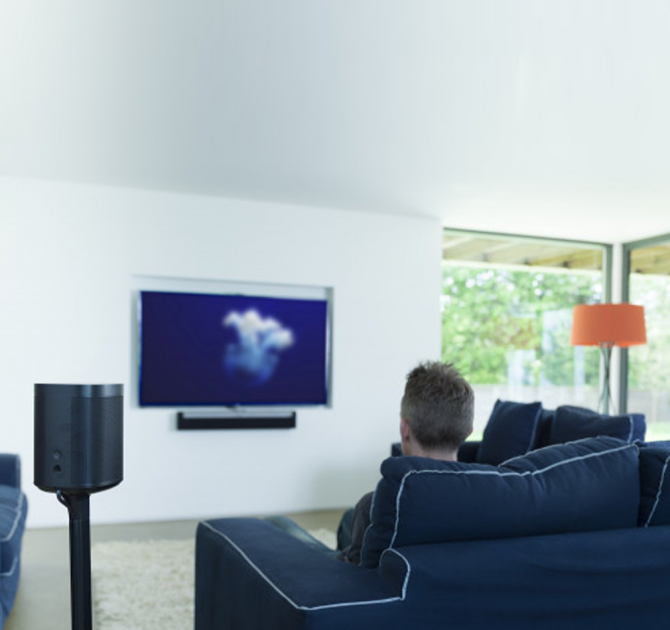 Flexson Adjustable Floor Stand One/Play1 with speaker and the view of a man from behind, sitting on a navy sofa watching a wall mounted television with an orange standard lamp in the corner of the room in front of a large double-aspect window.