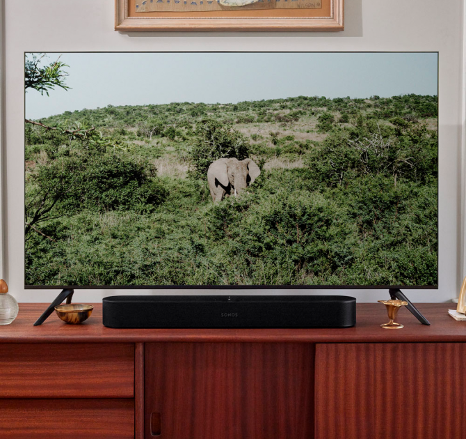 SONOS Beam (Gen 2) in black on top of a tv cabinet in wood.  The tv has a picture of an elephant in amongst trees and bushes.  The elephant is alone.