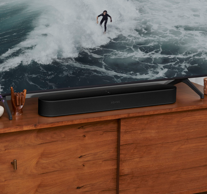 SONOS Beam (Gen 2) in black on top of a tv unit.  the tv has a picture of a surfer in a black wetsuit riding a wave.