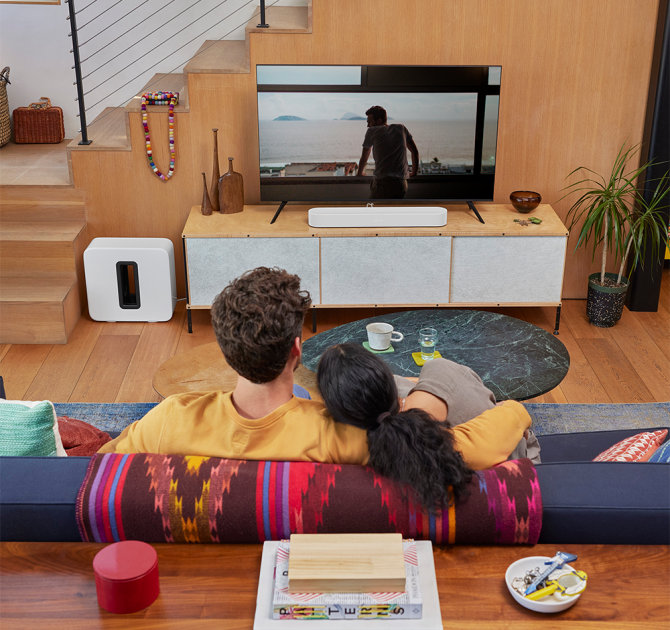 SONOS Beam (Gen 2) in white on top of a tv cabinet.  There is a Sonos Sub in white on the floor.  the tv is showing a man with his back to us looking out of a large window onto a very plain landscape.  there's a man and woman in the foreground with their back to us sitting close on a sofa.