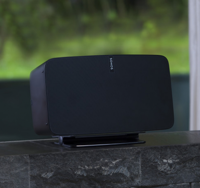 Flexson Desk Stand with a black Sonos Play 5 on it on a dark brick windowsill in front of a window.