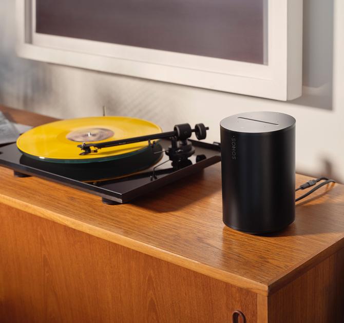 SONOS Era 100 Loudspeaker in black beside a black turntable playing a yellow record