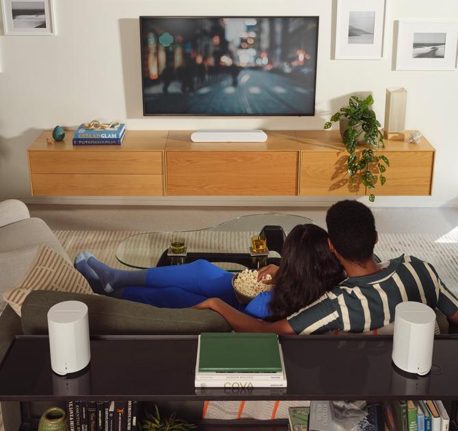 SONOS Era 100 Loudspeaker - a pair on a table in white behind a couple that are snuggling on a sofa watching tv