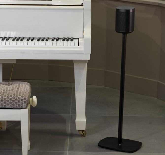 Flexson Floor Stand One/Play1 EU x1 in black with speaker next to a white piano with the piano stool partially visible.  In a room with a taupe tiled floor.
