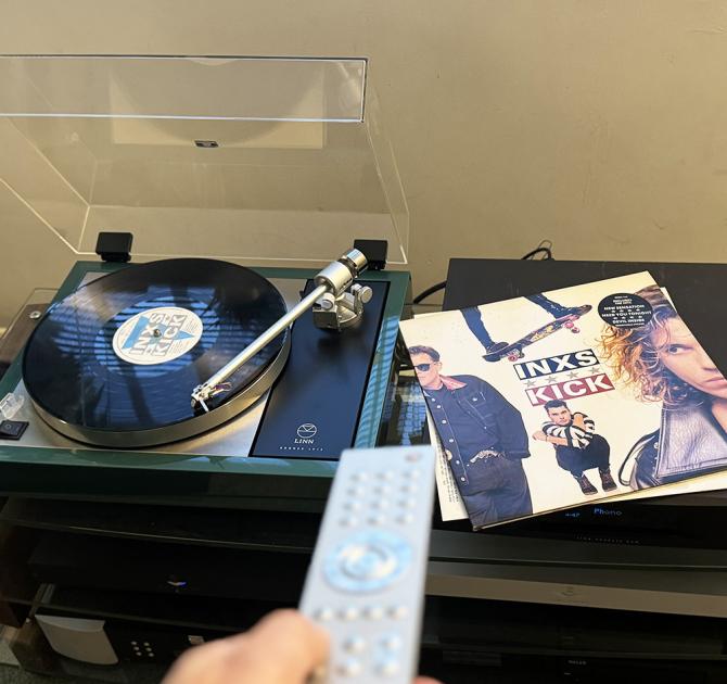 Linn Majik LP12 in a custom green colour.  It’s to the left of the image with an INXS vinyl album laying face up to the right. In the foreground a silver Linn remote control is pointing towards the turntable.