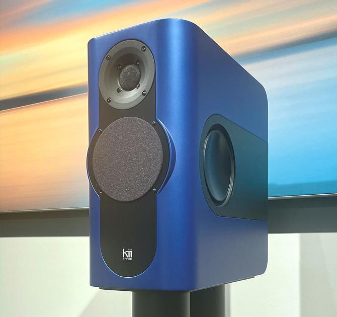 A blue rectangular speaker on a black stand with a piece of artwork in the background.