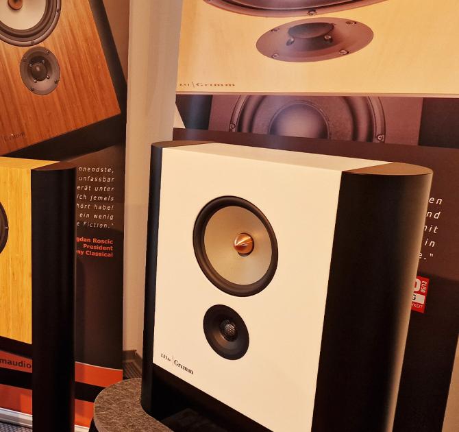 Grimm LS1be Loudspeaker in white on a table at the Munich High End Show in 2024.  there are images of speakers behind it and on the left of the image part of another speaker can be seen.