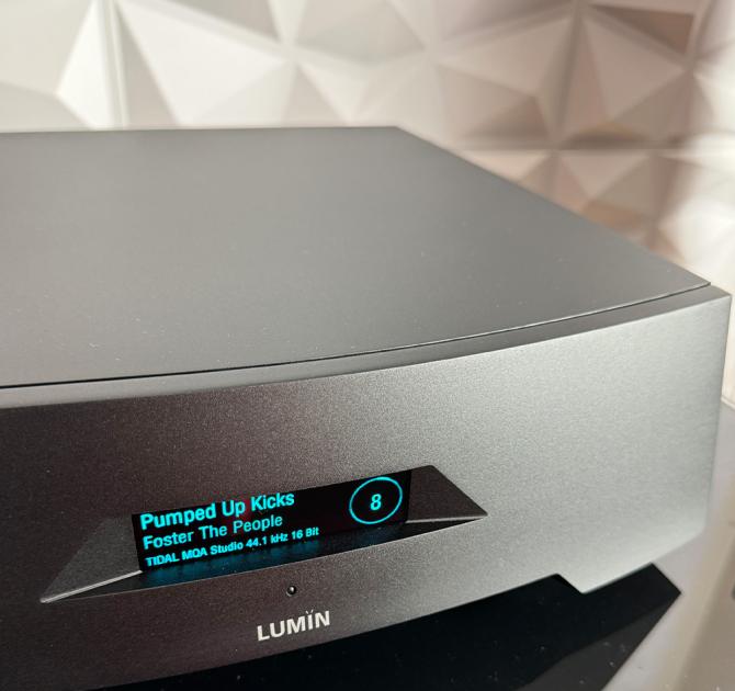 Lumin P1 in black with a geometric wall covering behind it.  The display shows the number "8" in a circle on the right.  On the left it shows "Pumped Up Kicks" then, under that, "Foster The People" then "Tidal MQA Studio 44.1 kHz 16 Bit"