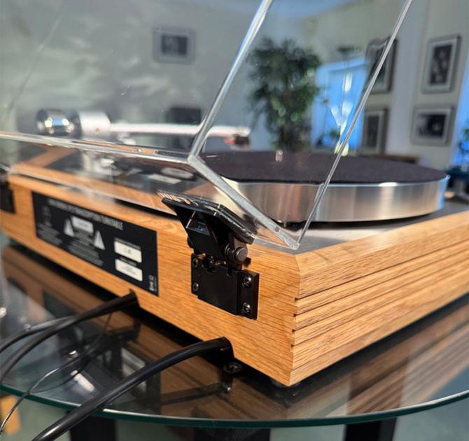 Linn Majik MM LP12 Fluted turntable in oak in the ripcaster showroom with sofas and speakers in the background along with other HiFi equipment.  This image shows a back corner with the hinge in focus