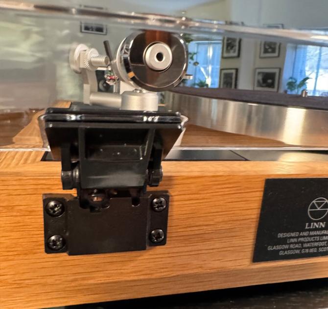 Linn Majik MM LP12 Fluted turntable in oak in the ripcaster showroom with sofas and speakers in the background along with other HiFi equipment.  This image is a close-up of a back hinge and the back of the tonearm.