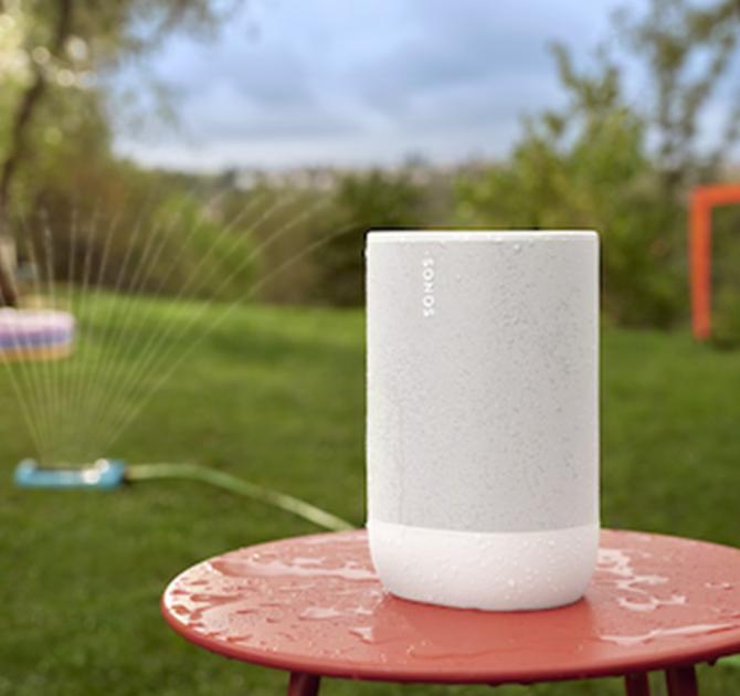 SONOS Move 2 Loudspeaker in white on a red table outside with a sprinkler.  The speaker has water droplets on it.